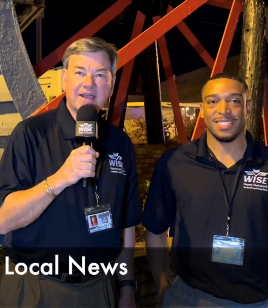 Two news reporters stand outdoors at night, one holding a microphone, both wearing black shirts with badges. A structure with red beams is in the background. The caption reads "Local News.