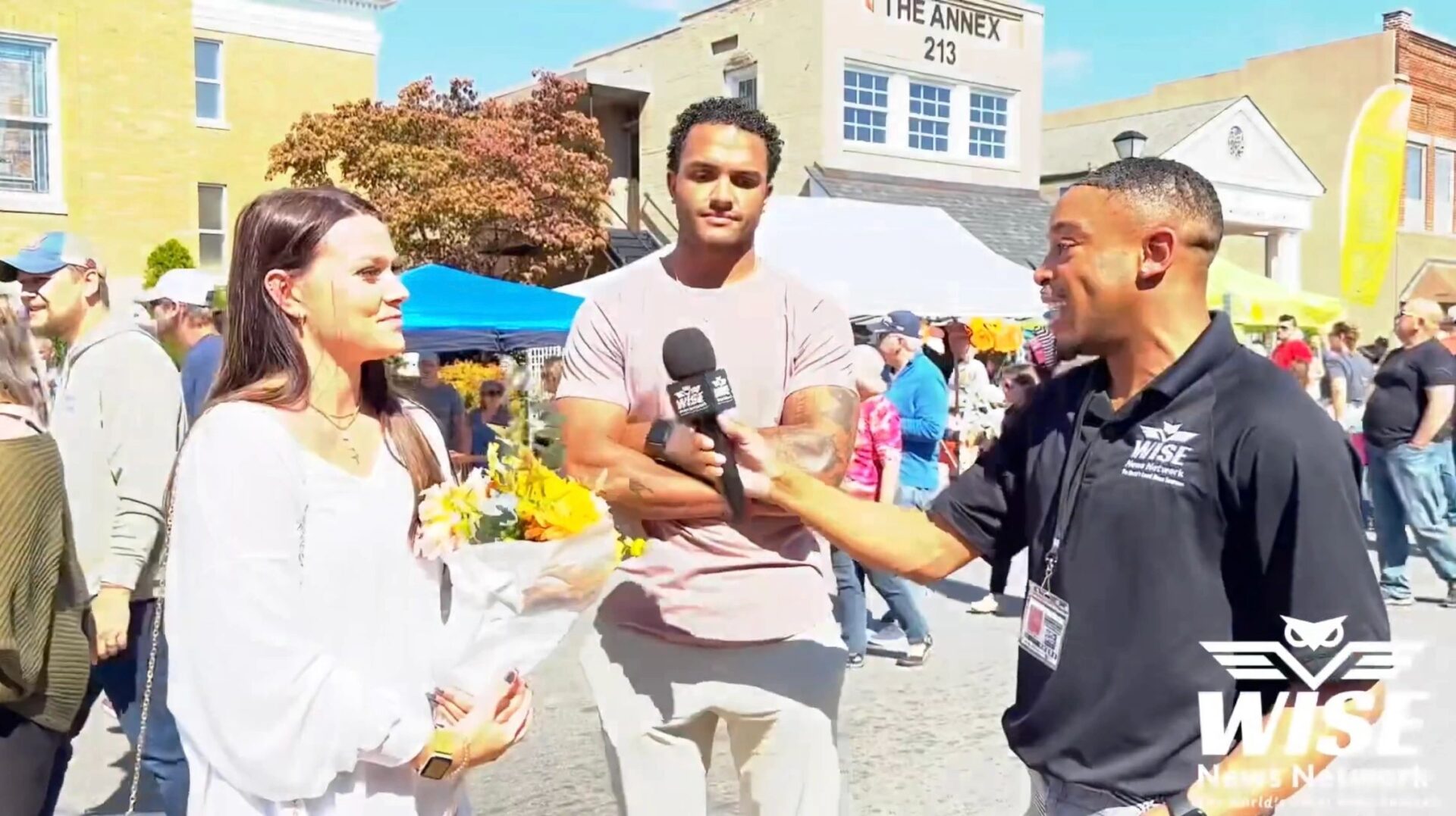 A person in a black shirt conducts an outdoor interview with a woman holding flowers and a man standing beside her. Other event attendees and tents are visible in the background.