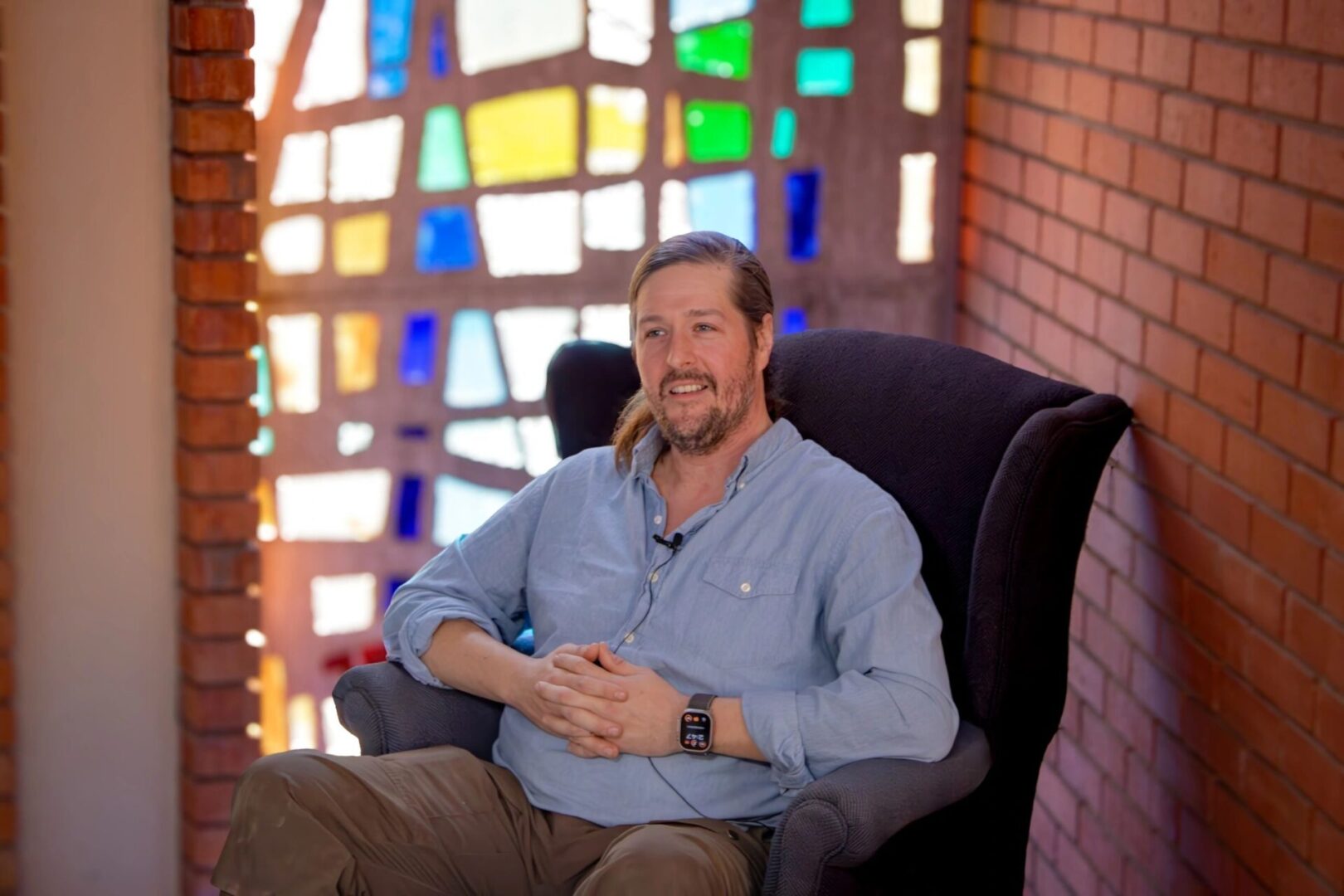 A person with long hair sits in a cushioned chair in front of a colorful stained glass window and brick wall, wearing a light blue shirt and a smartwatch.