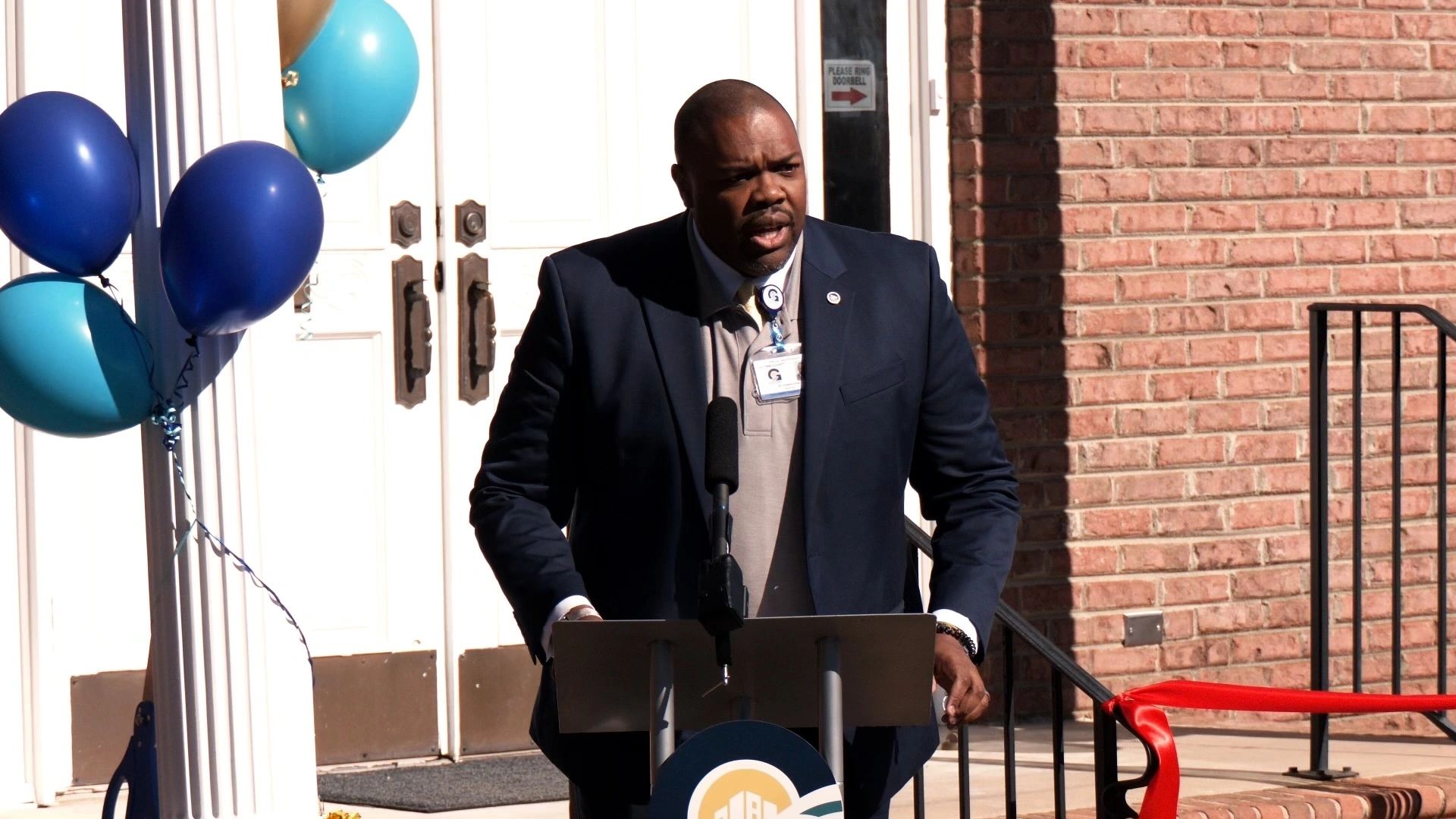 A man stands at a podium giving a speech outside a building. He is dressed in a dark suit. Balloons and a red ribbon are visible in the background.