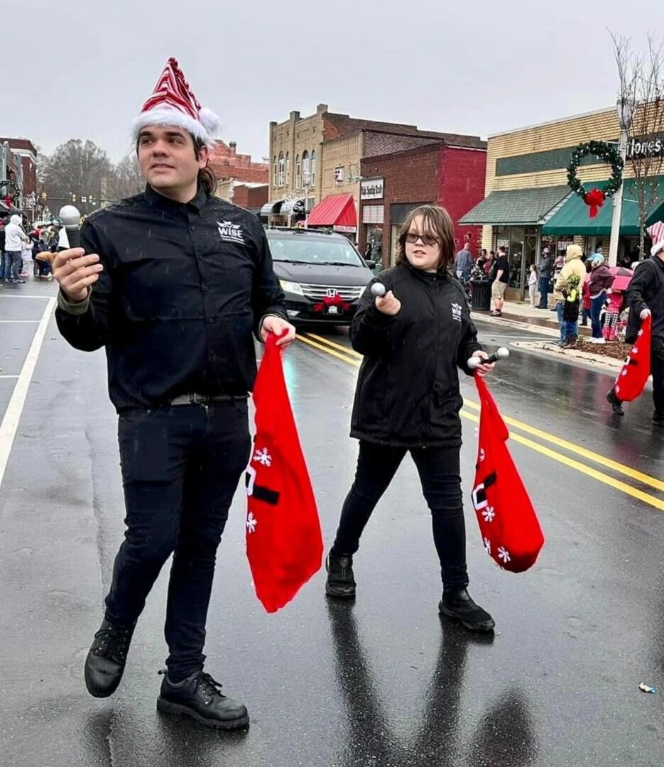 Two people walk in a parade, holding red bags and wearing holiday attire. They are on a street lined with small buildings and spectators. One wears a Santa hat.