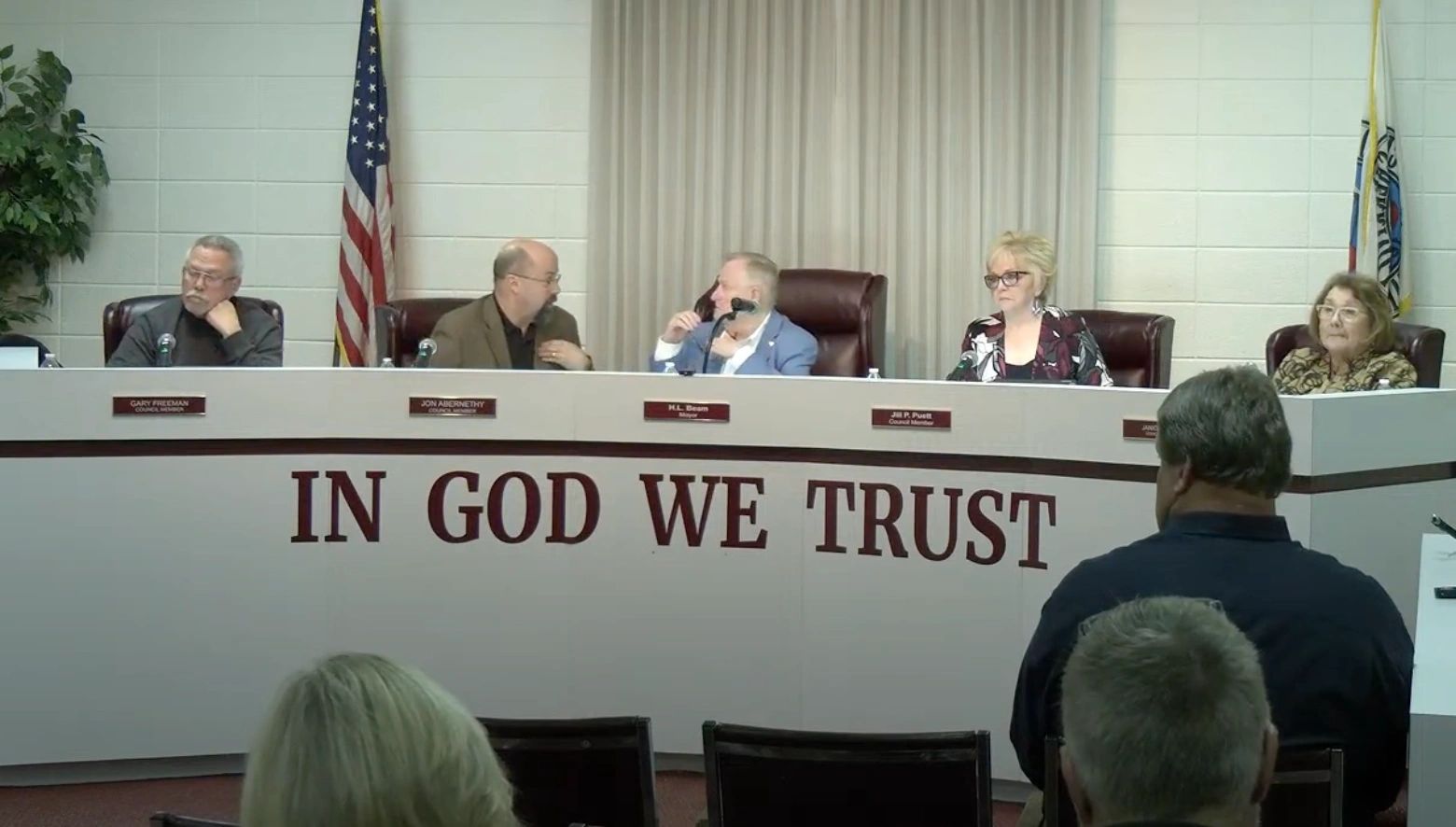 A panel of five individuals seated at a long table during a meeting, with "IN GOD WE TRUST" inscribed on the front; an audience member stands and addresses them.