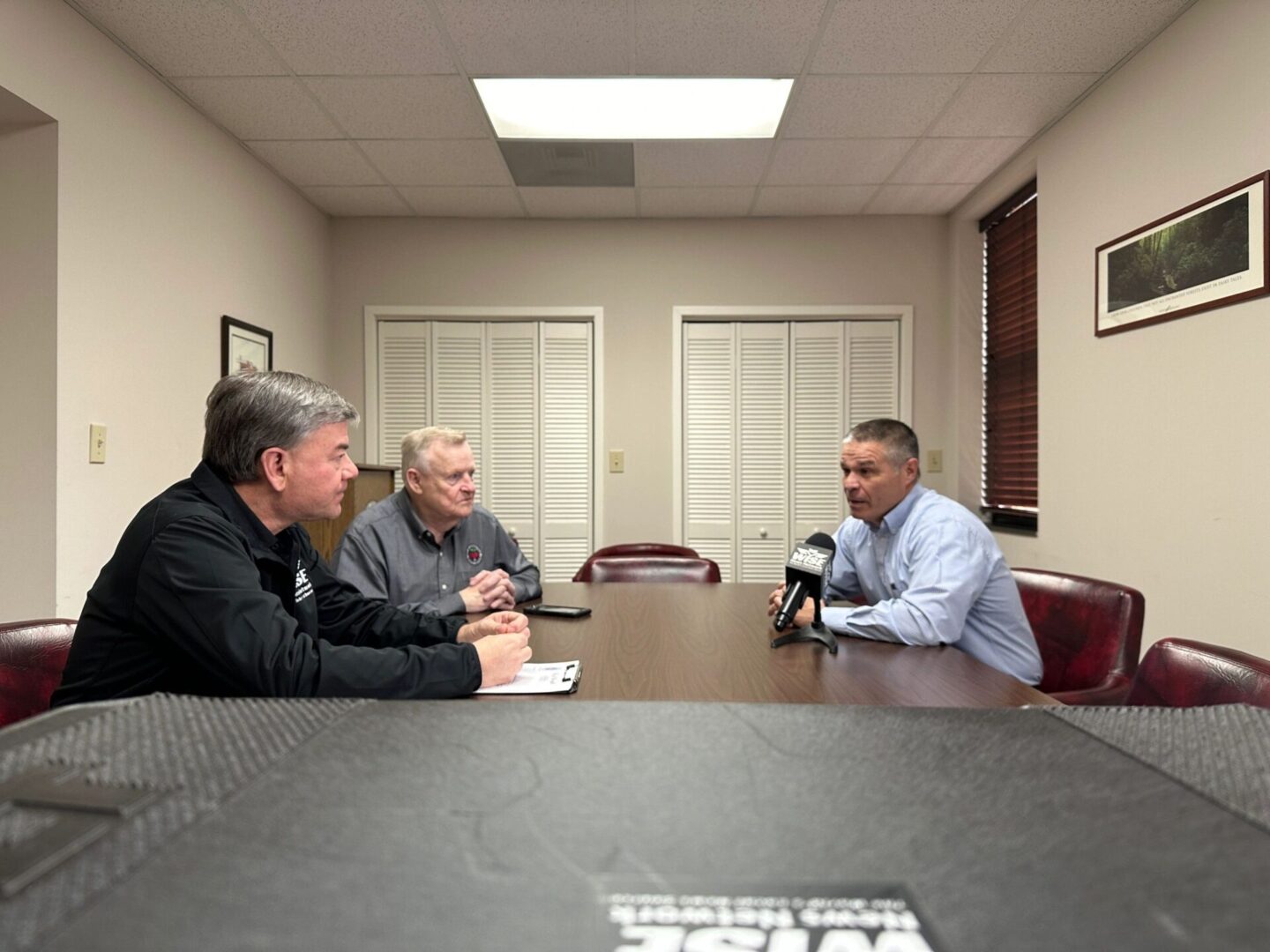 Three men are seated at a table in a conference room, engaged in a discussion. One man holds a microphone with the others listening attentively.