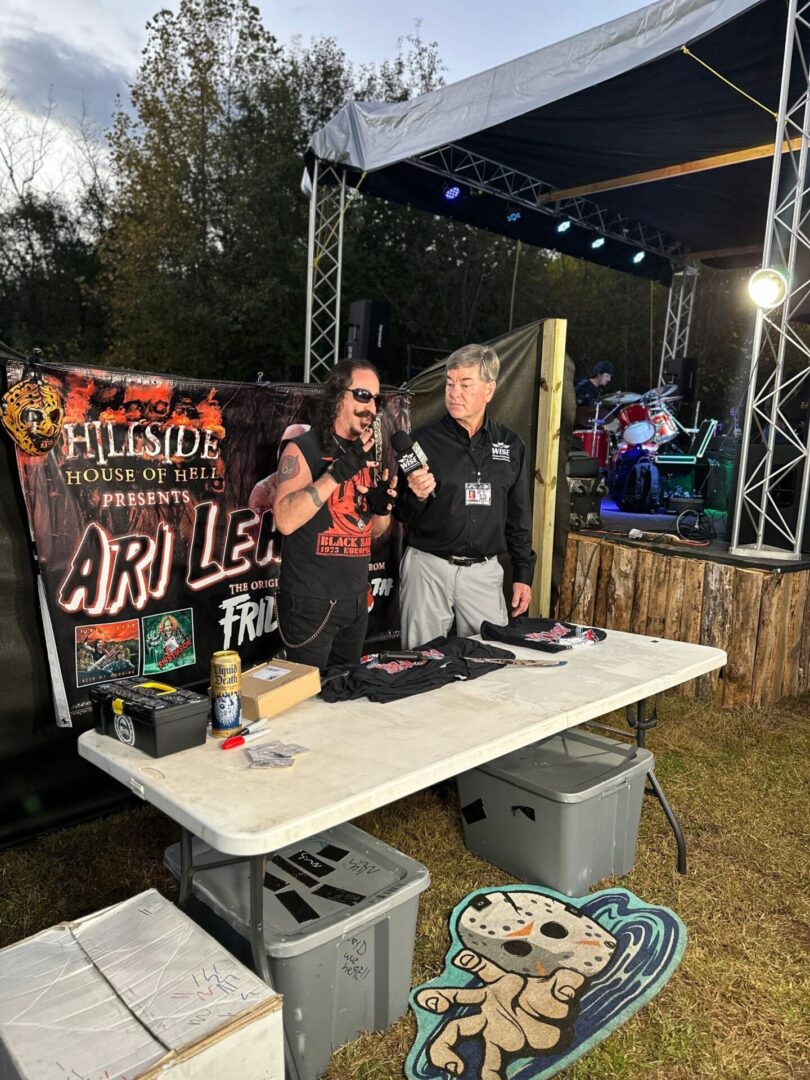 Two men stand at a merchandise table near a stage at an outdoor event. One man has long hair and sunglasses, the other has short hair and glasses. Various items are displayed on the table for sale.