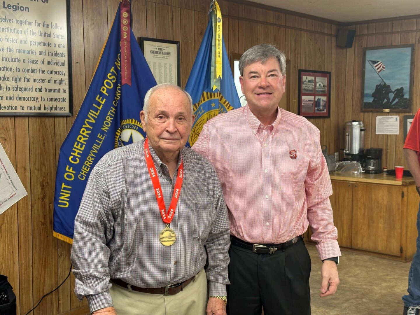 Two men stand together indoors. One man, wearing a medal, is older and dressed in a tan outfit, while the other man is younger and dressed in a red shirt and black trousers. Flags are in the background.