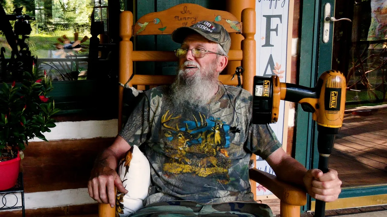 A man with a beard wearing a camouflage T-shirt and cap sits in a wooden rocking chair on a porch, holding a power drill.