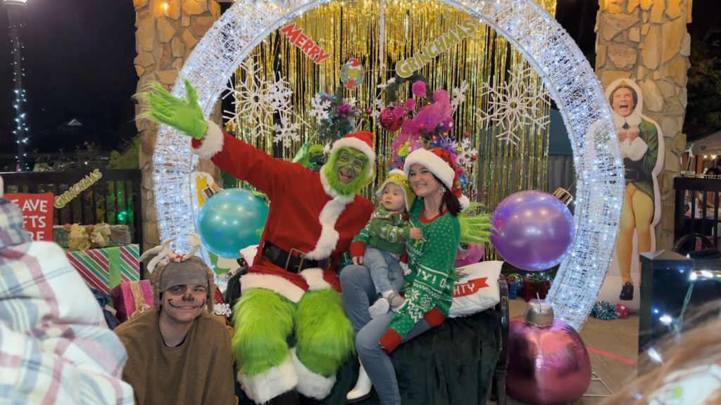 A man in a Grinch outfit with a red santa suit posing with a woman and her newborn at a festive scene. 