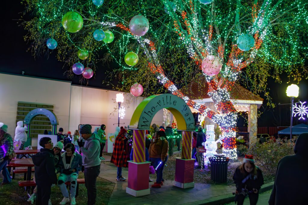 A crowd appearing in a park decorated with lights, with an arch reading "Who-Ville Park". 