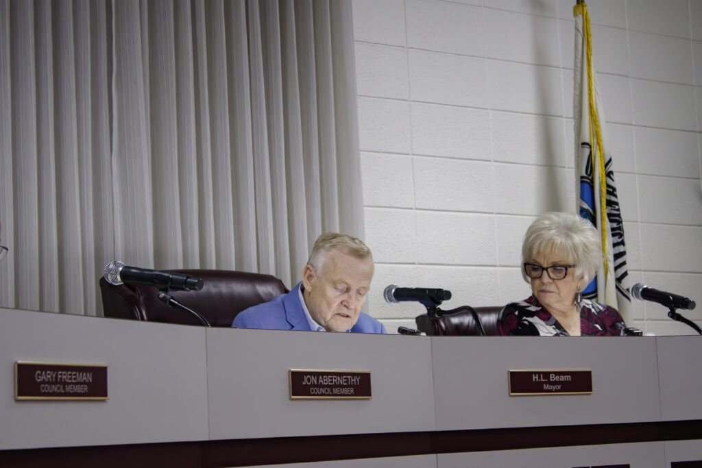 A man and woman sitting behind a counter, the man is wearing a blue shirt and speaking into a microphone and the woman is wearing a black shirt and glasses. 