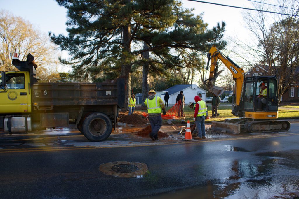 An image showing 7 men working on a hole in the road that is leaking water. There is also a dump truck and excavator in use. 