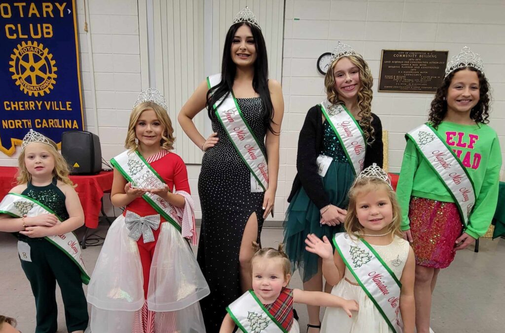 7 women of various ages who are all wearing green and white sashes and are otherwise dressed in a christmas theme.