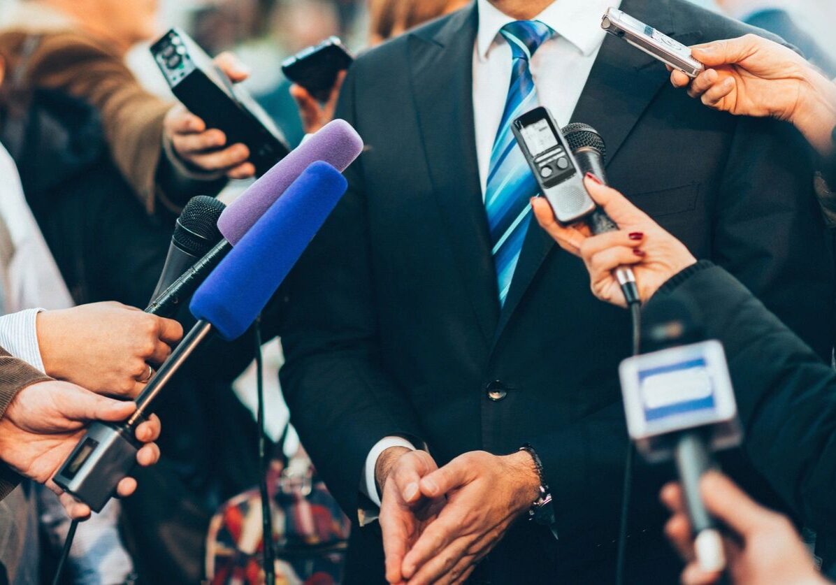 A person in a suit is surrounded by journalists holding microphones and recording devices during an interview.