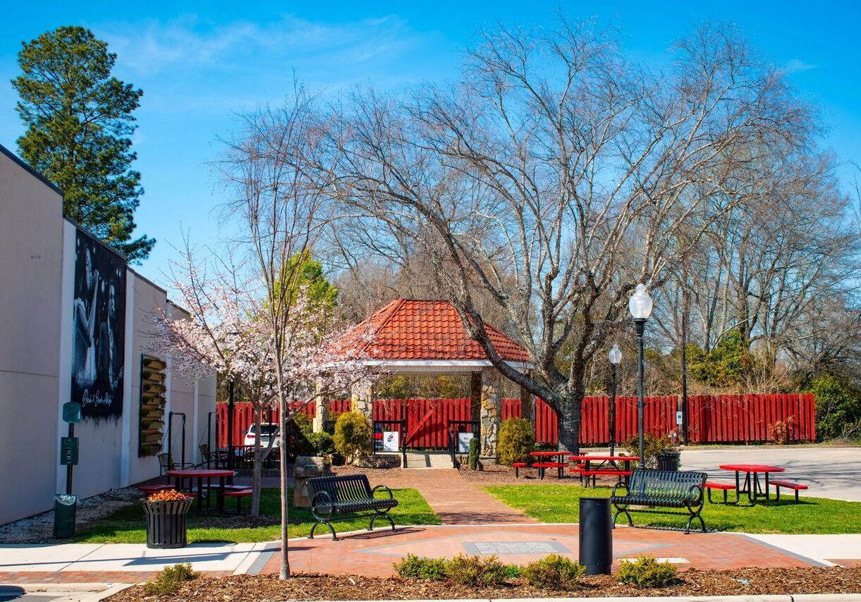 A small park with red picnic tables, green grass, and blossoming trees. A building with a red roof is in the background, and a mural is on the left wall. The sky is clear and blue.