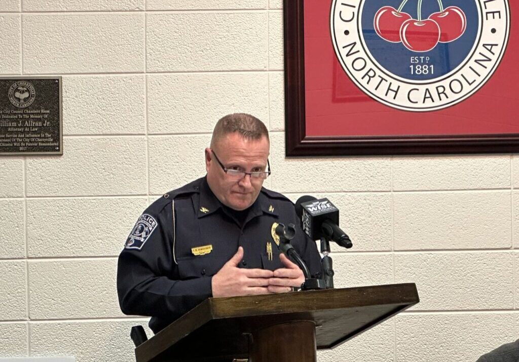 A police officer in uniform stands at a podium, speaking into microphones, with a city emblem on the wall behind him.