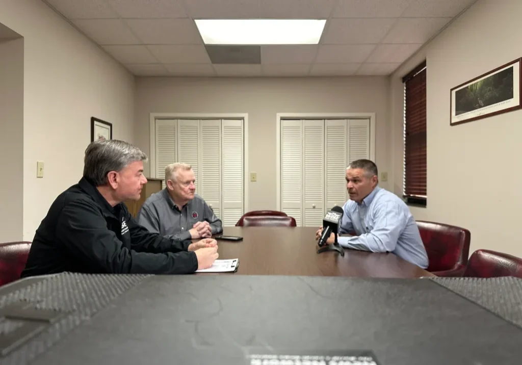 Three men sit around a conference table, engaging in a discussion. One man holds a microphone at the center. The room has minimal decoration with closed blinds and framed art on the walls.