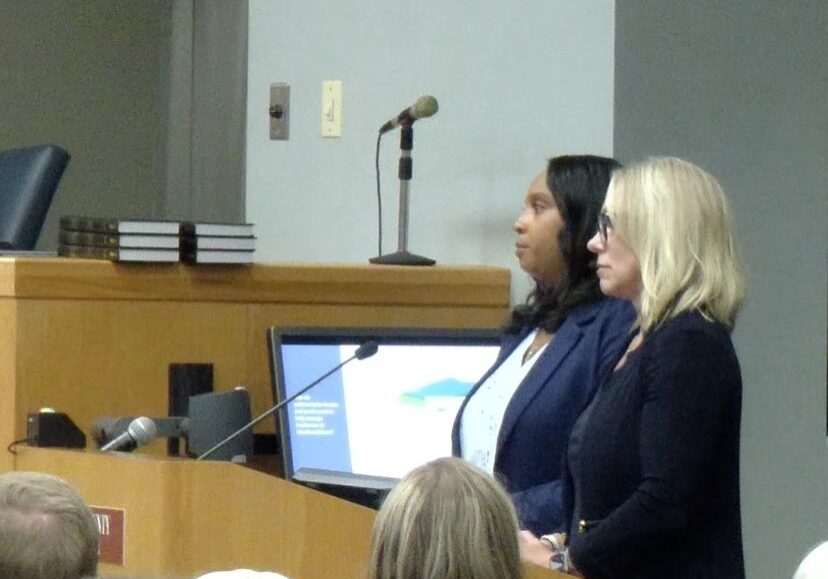 Two women stand at a podium giving a presentation in front of an audience; a microphone and a monitor displaying a slide are visible. The setting appears to be a formal meeting or conference.