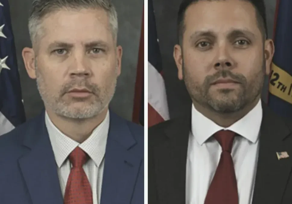 Two men in formal attire pose for portraits. Both wear suits with ties; one in a blue suit with a red tie, the other in a black suit with a red tie. Flags are visible in the background.