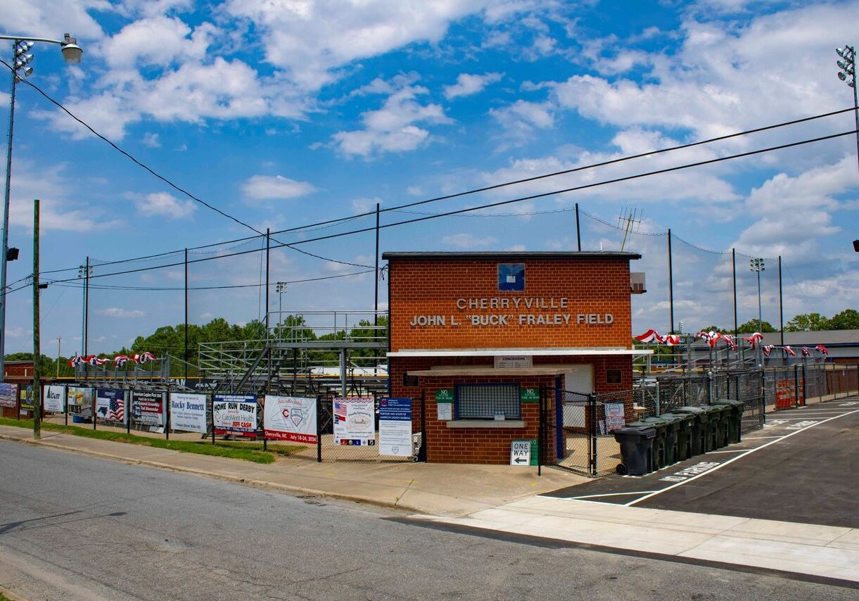 A brick building with "Cherryville, John L. Buck Fraley Field" signage, surrounded by fences, banners, and parked trash bins under a partly cloudy sky.