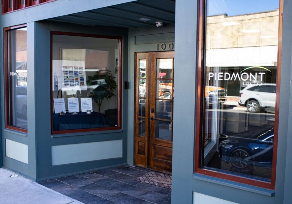 Street view of the entrance to a store with a wooden door and large windows displaying the name "Piedmont." Reflections of cars and buildings are visible in the glass.