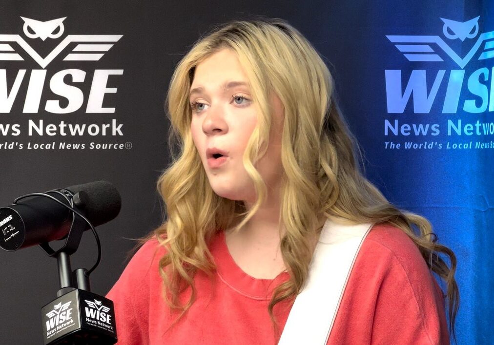 A person with long blonde hair in a red top speaking or singing into a microphone at a WISE News Network event.