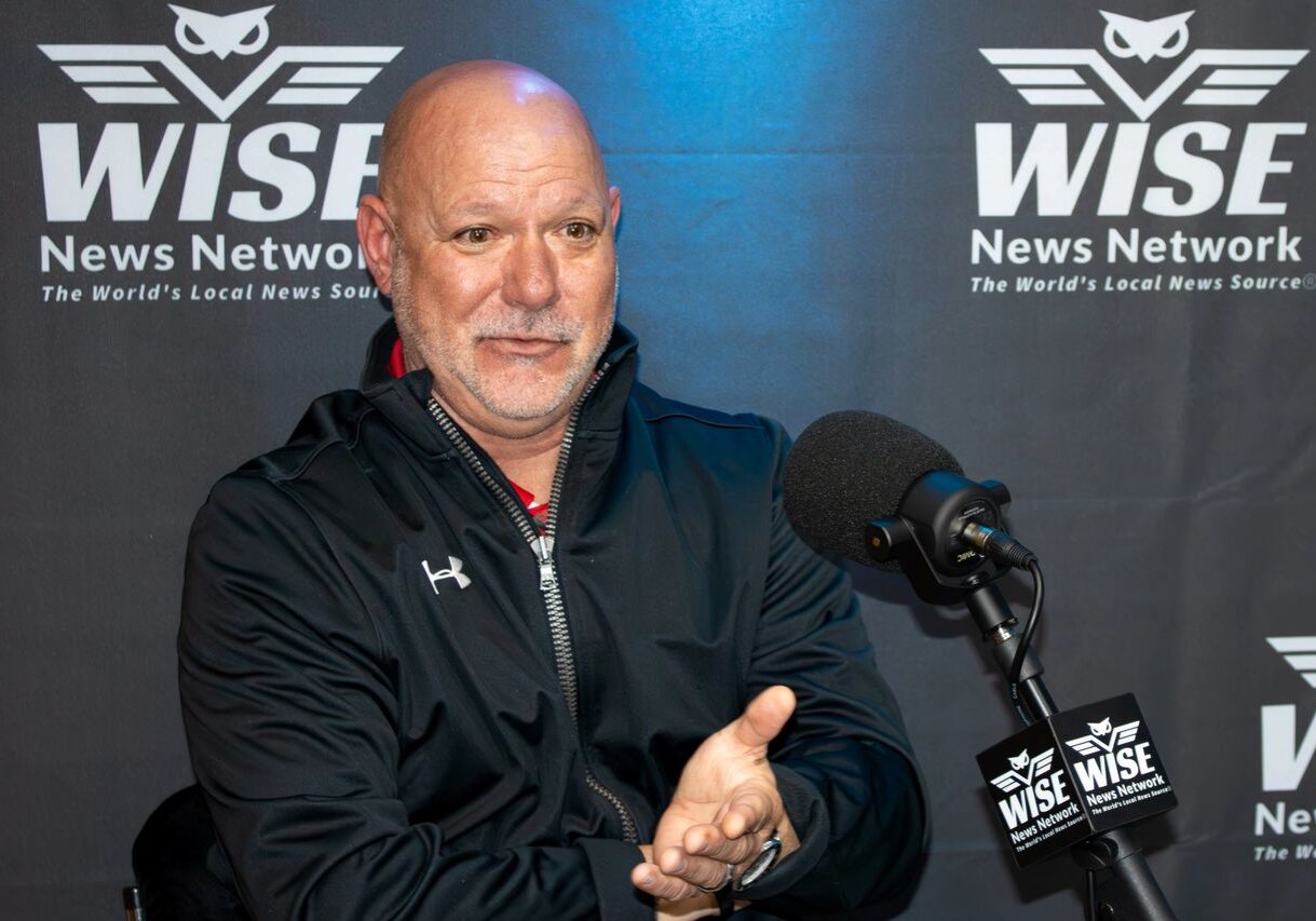 A man with a bald head and goatee, wearing a black jacket, speaks into a microphone in front of a WISE News Network backdrop.