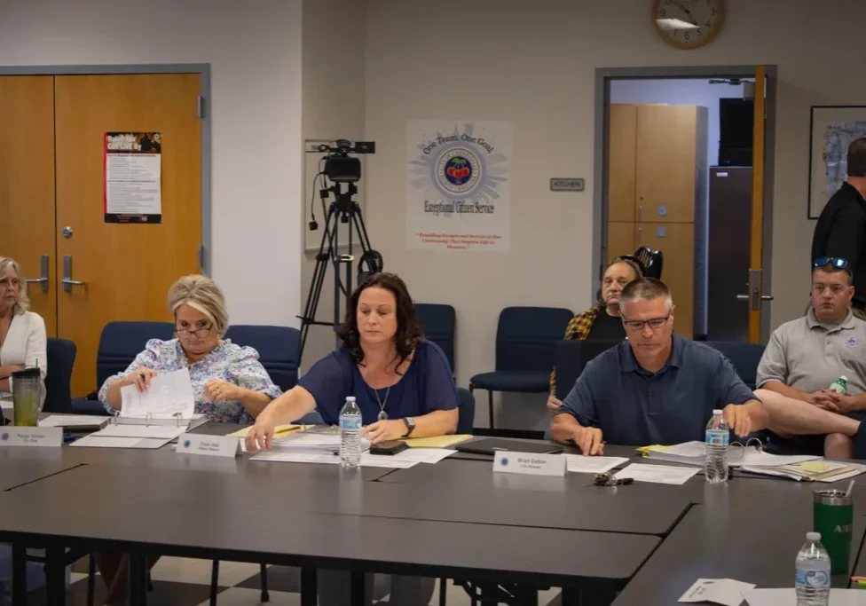 Several individuals are seated at a conference table with documents and nameplates during a meeting. A camera and poster are visible in the background.