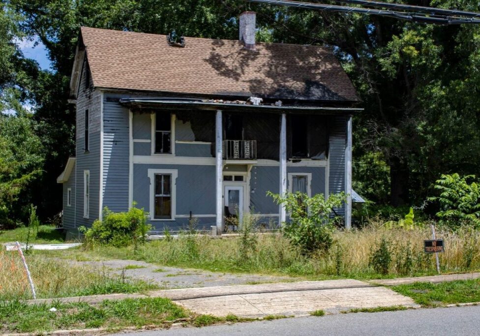 A dilapidated two-story house with faded blue paint, boarded-up windows, overgrown yard, and a "For Sale" sign out front. The house has two large white pillars supporting a small upper balcony.