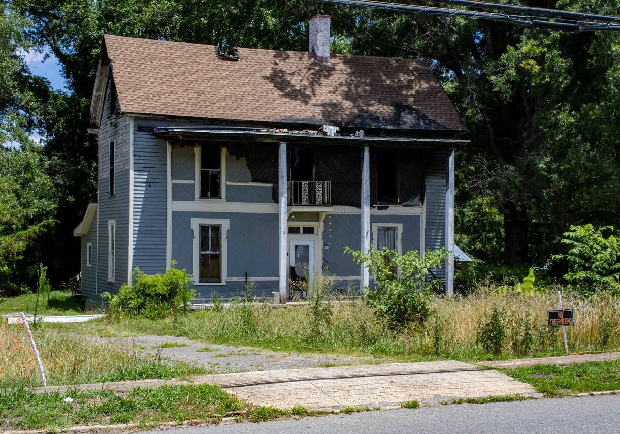 A dilapidated two-story house with faded blue paint, boarded-up windows, overgrown yard, and a "For Sale" sign out front. The house has two large white pillars supporting a small upper balcony.