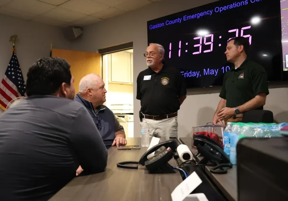 A group of four men in a meeting room with an American flag and a large digital clock displaying the date and time. They are engaged in discussion, with emergency operation equipment on the table.