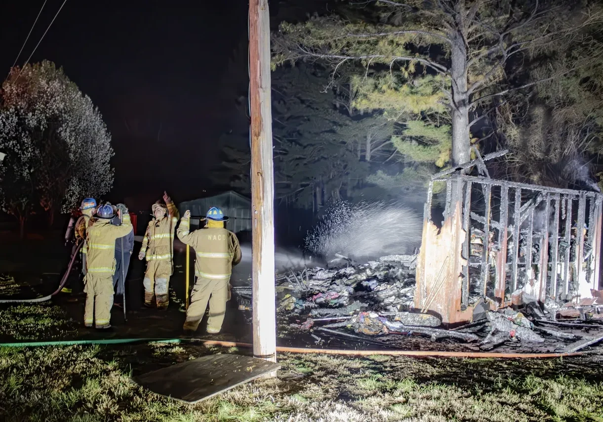 Firefighters work at the scene of a night-time fire, extinguishing flames amidst the charred remains of a structure next to a partially burned tree.