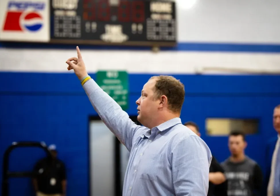 A man in a light blue shirt points upwards, standing inside a gymnasium with a scoreboard and other individuals in the background.