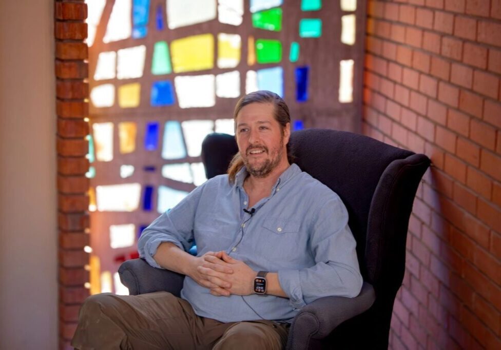 A person wearing a light blue shirt sits in a black armchair, with colorful stained glass and brick walls in the background. They are smiling, and a microphone is clipped to their shirt.