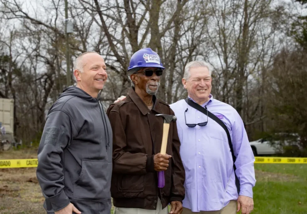 Three men stand together outdoors; one holds a hammer and wears a purple hard hat, posed in front of a wooded area with caution tape in the background.