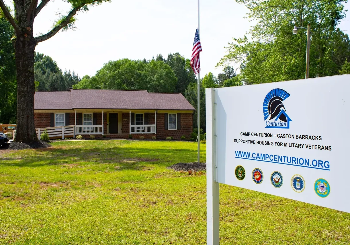 A single-story brick building with a sign in the foreground that reads "Camp Centurion - Gaston Barracks: Supportive Housing for Military Veterans" and the website www.campcenturion.org. An American flag is on a pole in the yard.