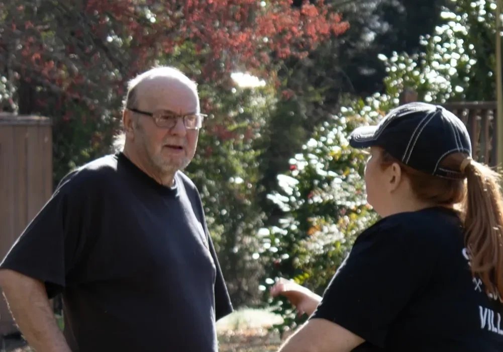 Two people engage in a conversation outdoors. The man on the left wears glasses and a dark shirt, while the woman on the right, with her hair in a ponytail and a dark cap, also wears a dark shirt. Trees are in the background.