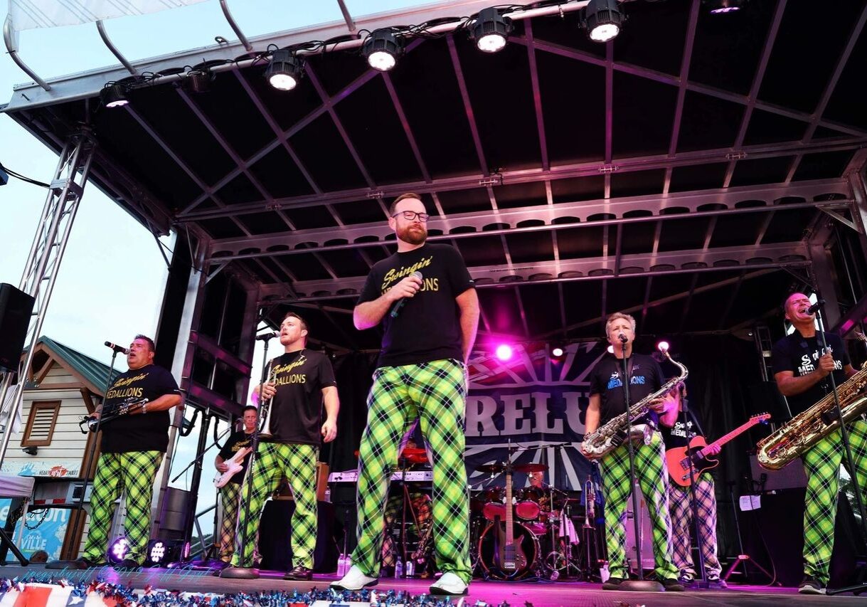 A band performs on an outdoor stage, wearing matching green plaid pants and black t-shirts. The stage is lit with purple lights and various musical instruments are visible.