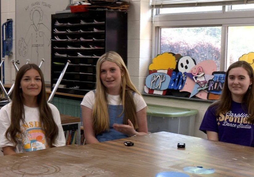 Three young people sit at a table in a classroom. The table has some scattered objects, and chairs are stacked on desks in the background. Colorful decorations are visible on the window sill.