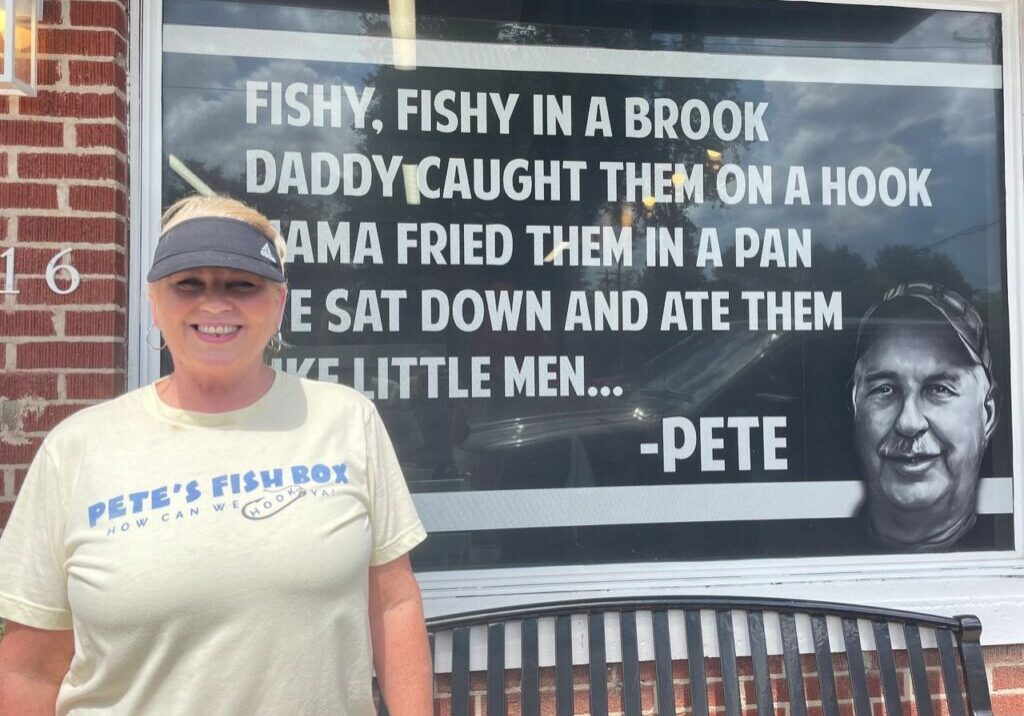 A woman in a "Pete's Fish Box" shirt stands in front of a window display featuring a fish-themed quote and an image of a man named Pete.