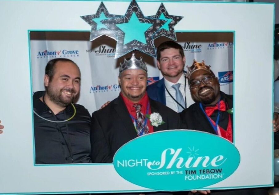 Four men smiling and wearing crowns pose behind a "Night to Shine" frame sponsored by the Tim Tebow Foundation. Three are dressed in suits, and one is casually dressed.