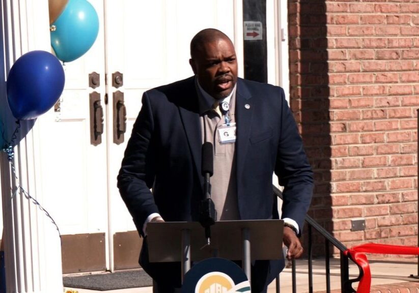 A man stands at a podium with microphones, speaking outside a brick building. Blue and gold balloons are on the left, and a red ribbon is visible on the right.