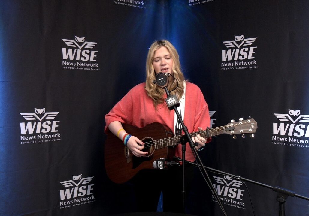 A person is playing an acoustic guitar and singing into a microphone in front of a backdrop with logos for "WISE News Network.