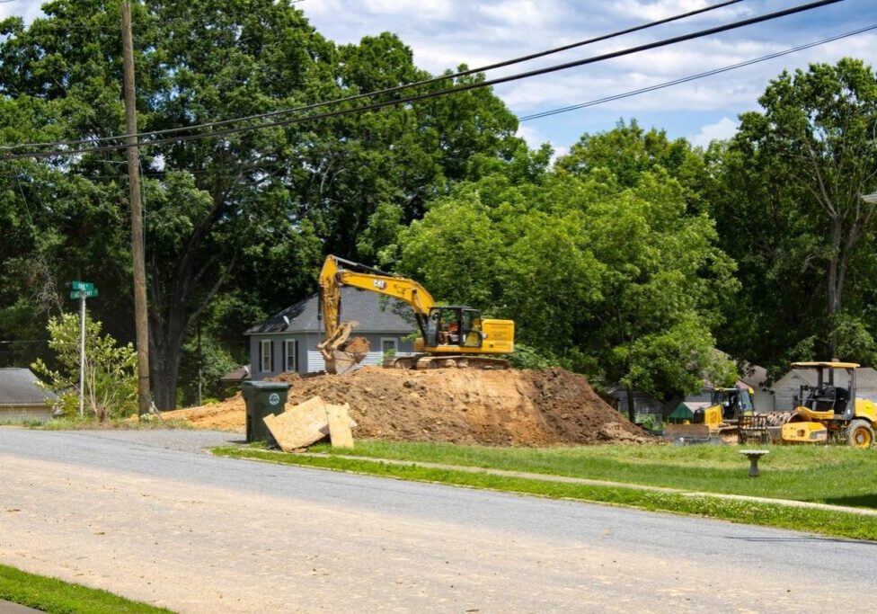 Construction site with an excavator on a mound of dirt, surrounded by trees and residential buildings. A road and a bulldozer are in the foreground.