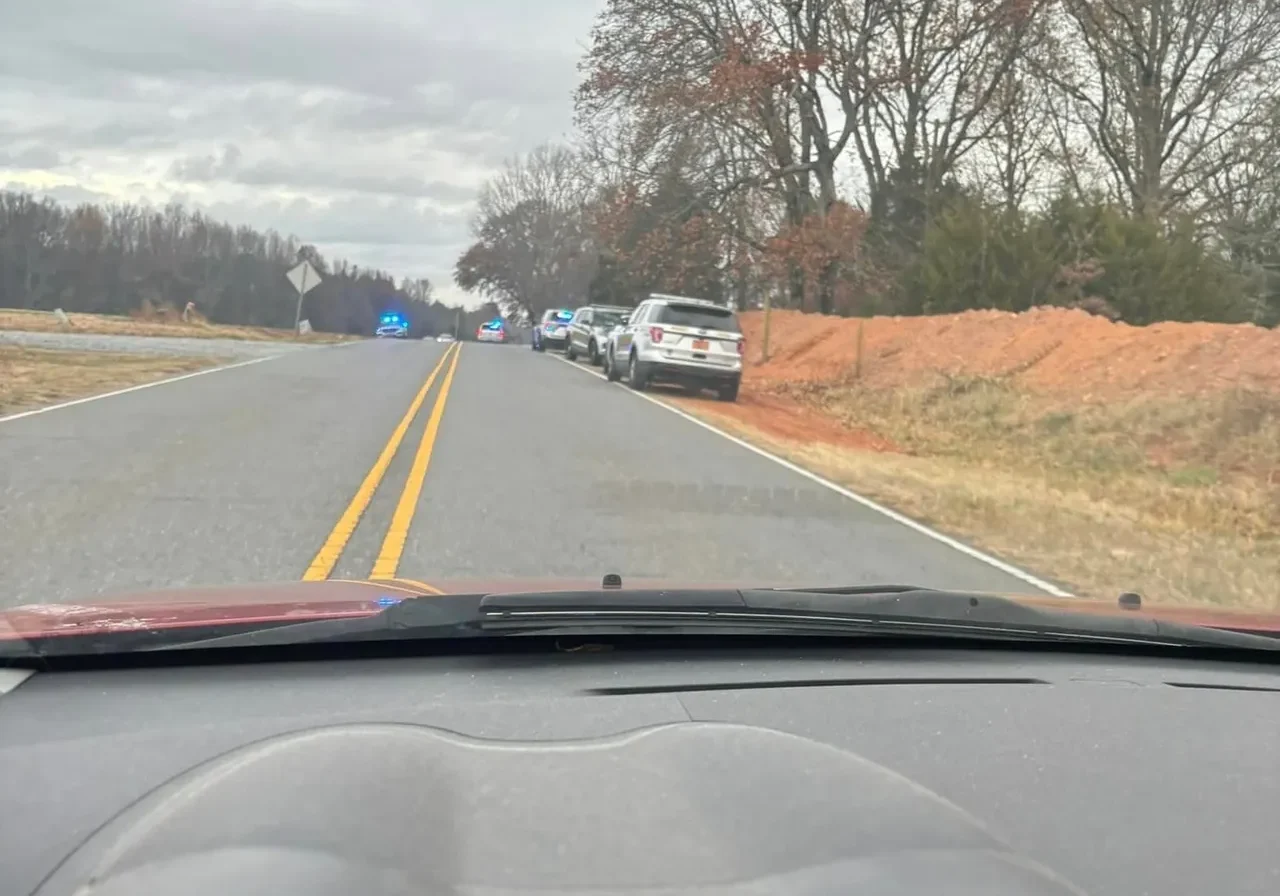 Several cars and police vehicles with flashing lights are parked on the side of a rural road lined with trees. The photo is taken from inside a car driving toward the scene.