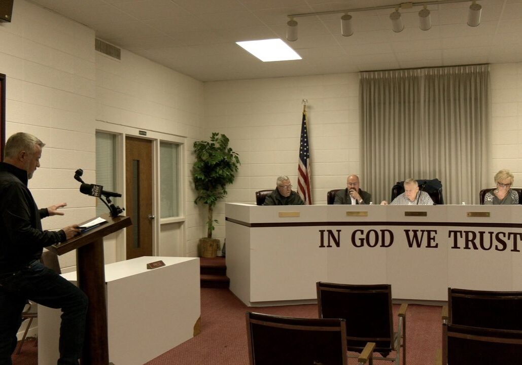 A man at a podium addresses four seated officials in a conference room. A wall sign reads "In God We Trust." An American flag is in the background.