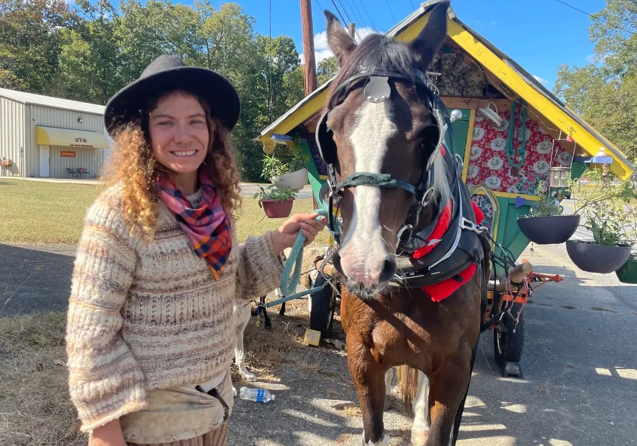 A woman in a hat stands next to a horse harnessed to a small, colorful wagon on a sunny day.