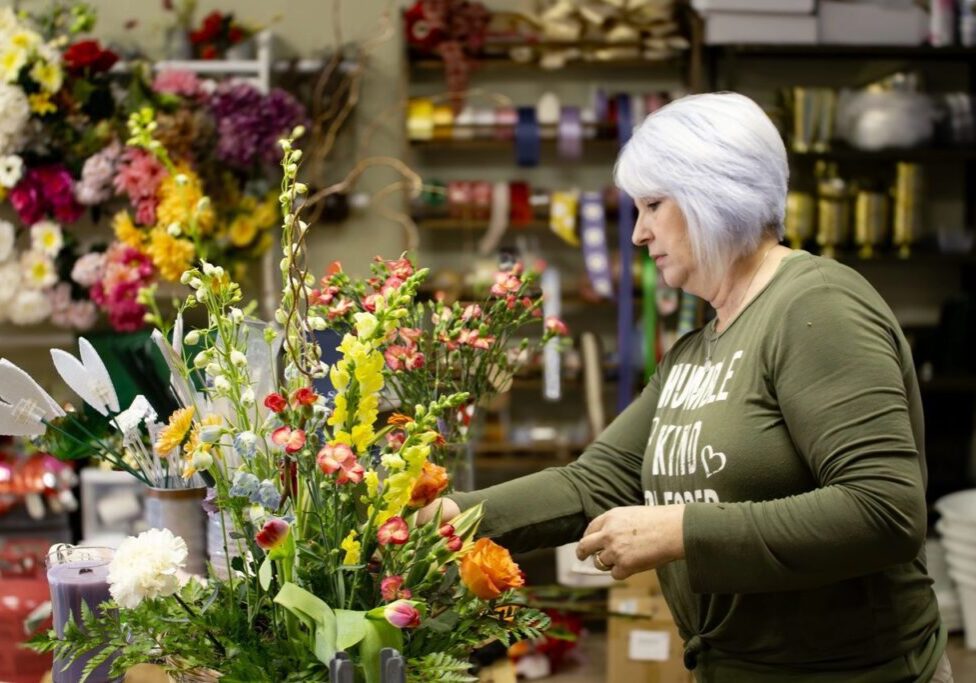 A person with short white hair arranges a colorful flower bouquet in a floral shop with various supplies and flowers in the background.