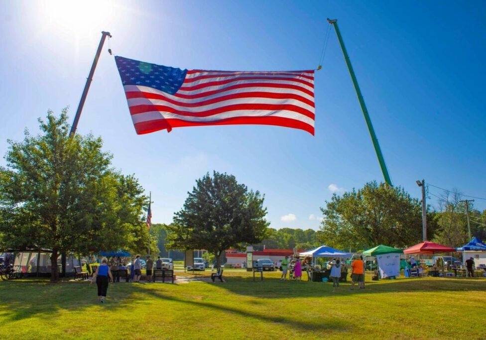 A large American flag is suspended between two cranes at an outdoor event with people gathering under tents and trees in a sunny park setting.