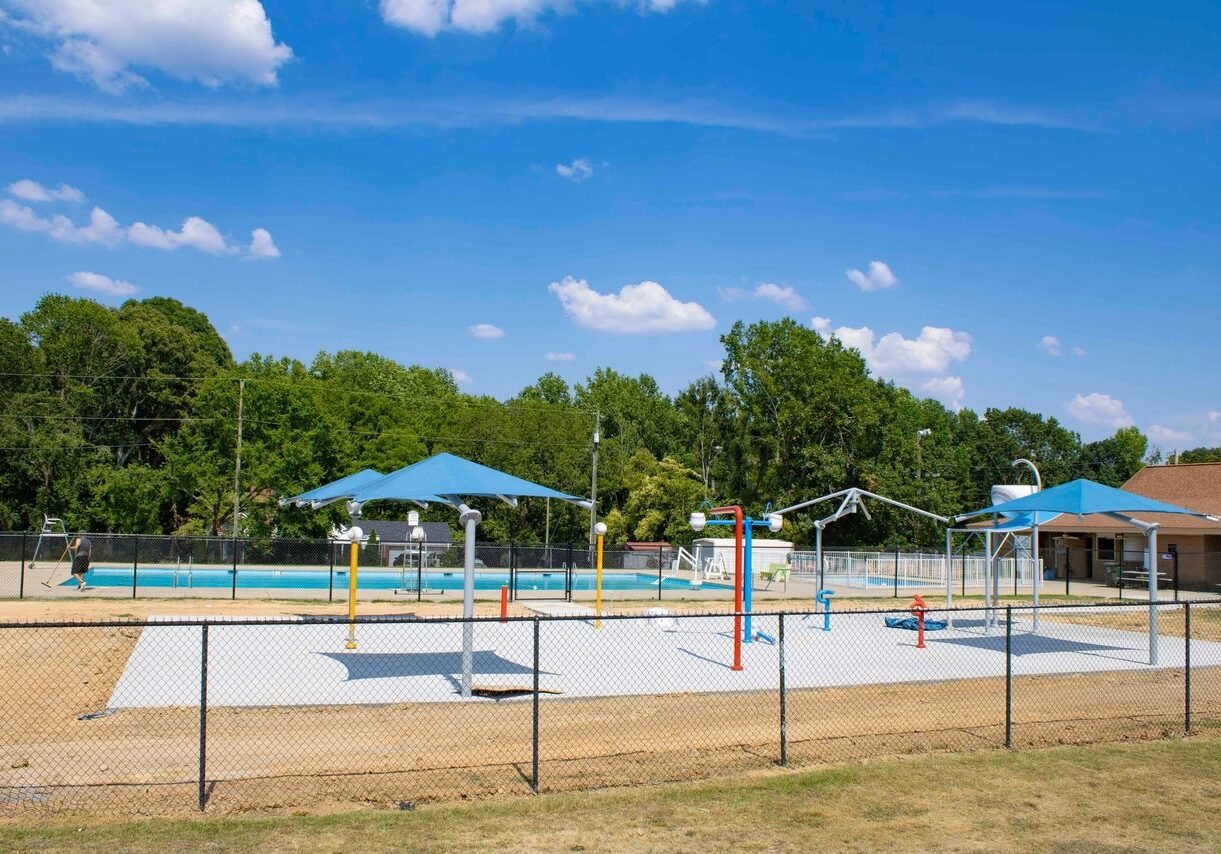 A fenced-off public pool area with a splash pad, shade structures, and adjacent building under a partly cloudy sky.