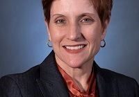 A person with short hair, wearing a dark blazer and a reddish-orange blouse, smiles for a professional portrait against a blue background.