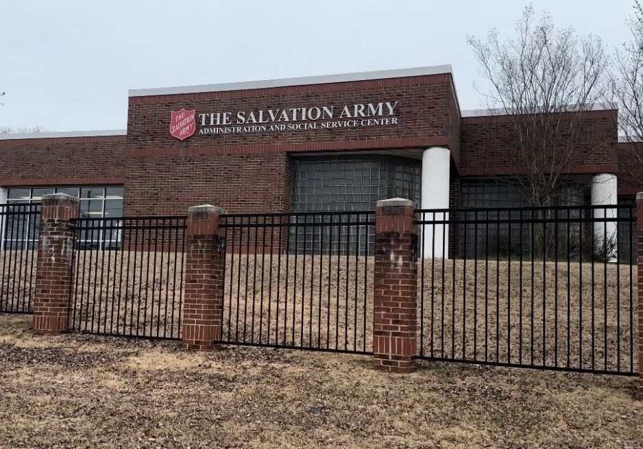 A brick building with "The Salvation Army Administration and Social Service Center" sign, behind a black metal fence and surrounded by patches of grass and trees.